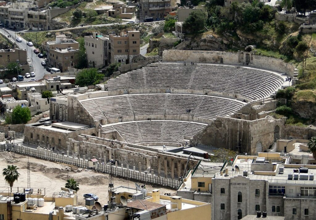 Ancient stone amphitheater with curved, tiered seating surrounded by town houses and hillsides.