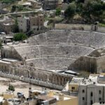 Ancient stone amphitheater with curved, tiered seating surrounded by town houses and hillsides.